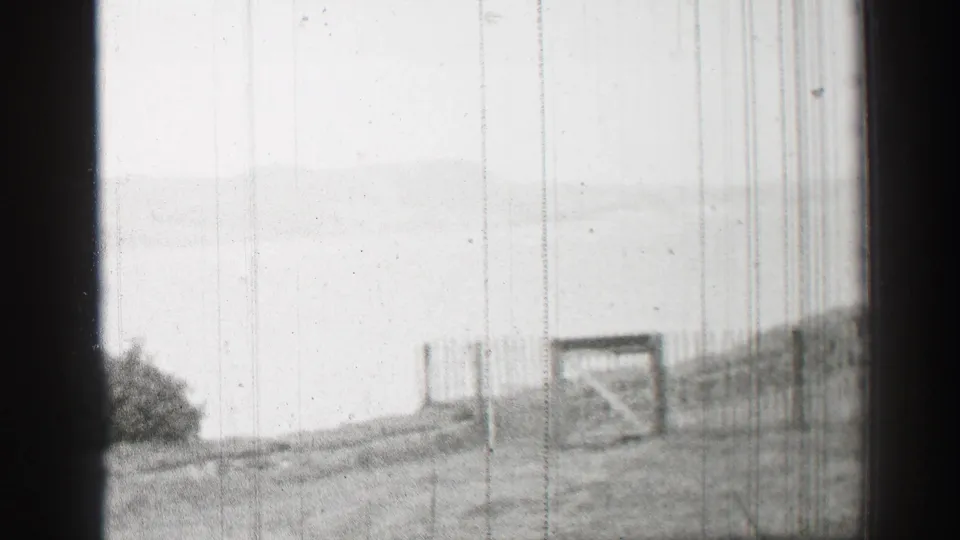 1952 United Kingdom 8mm frame looking across a closed gate, wire fencing, grassy slope, and a pale distant horizon that may be water.
