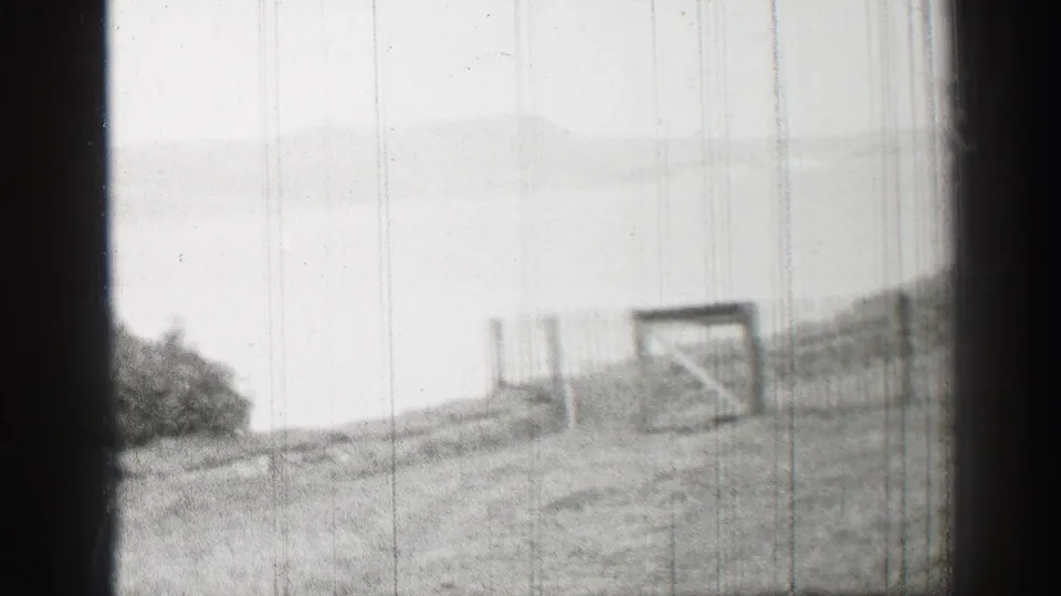 1952 United Kingdom home-movie frame showing a soft black-and-white view across a grassy lakeside slope toward water and distant hills.