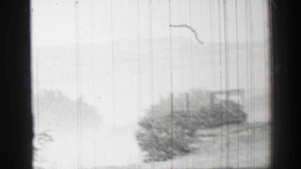 1952 United Kingdom home-movie frame with pale water, distant hills, and a dark bush or tree mass along the right shoreline.