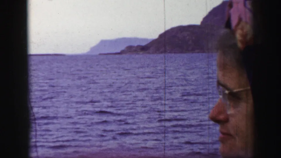 Older woman in glasses and a patterned headscarf sits in profile beside a window looking toward a rocky island across the water.