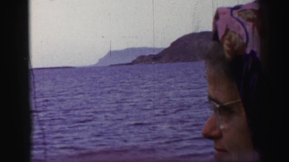 1952 8mm frame of a woman in glasses and headscarf near a ship window, with open sea and a distant mountainous coastline visible.