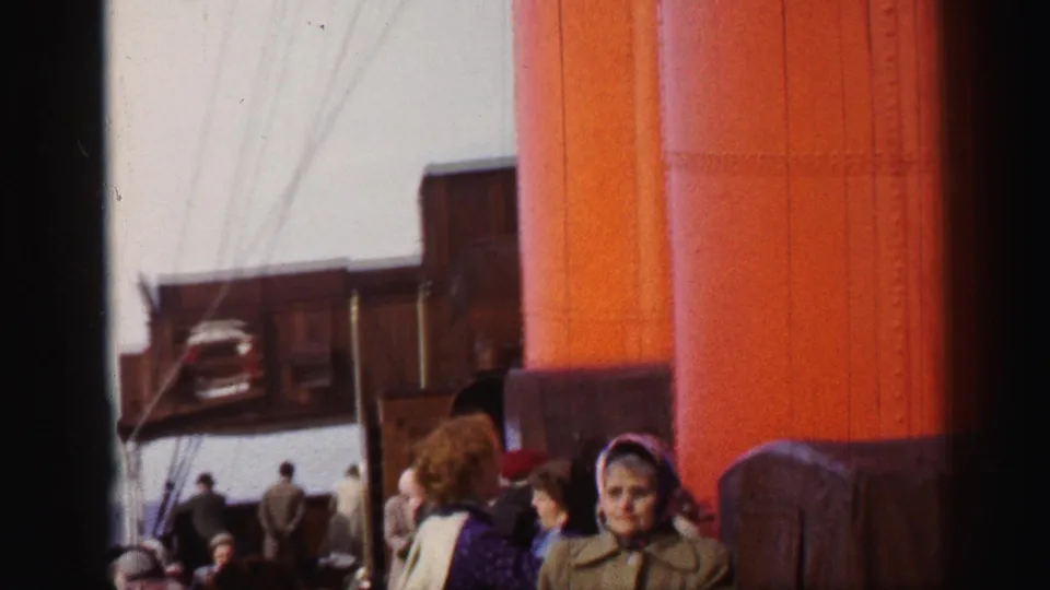 1952 8mm frame of passengers and luggage on a ship deck, with the orange smokestack dominating the right side.