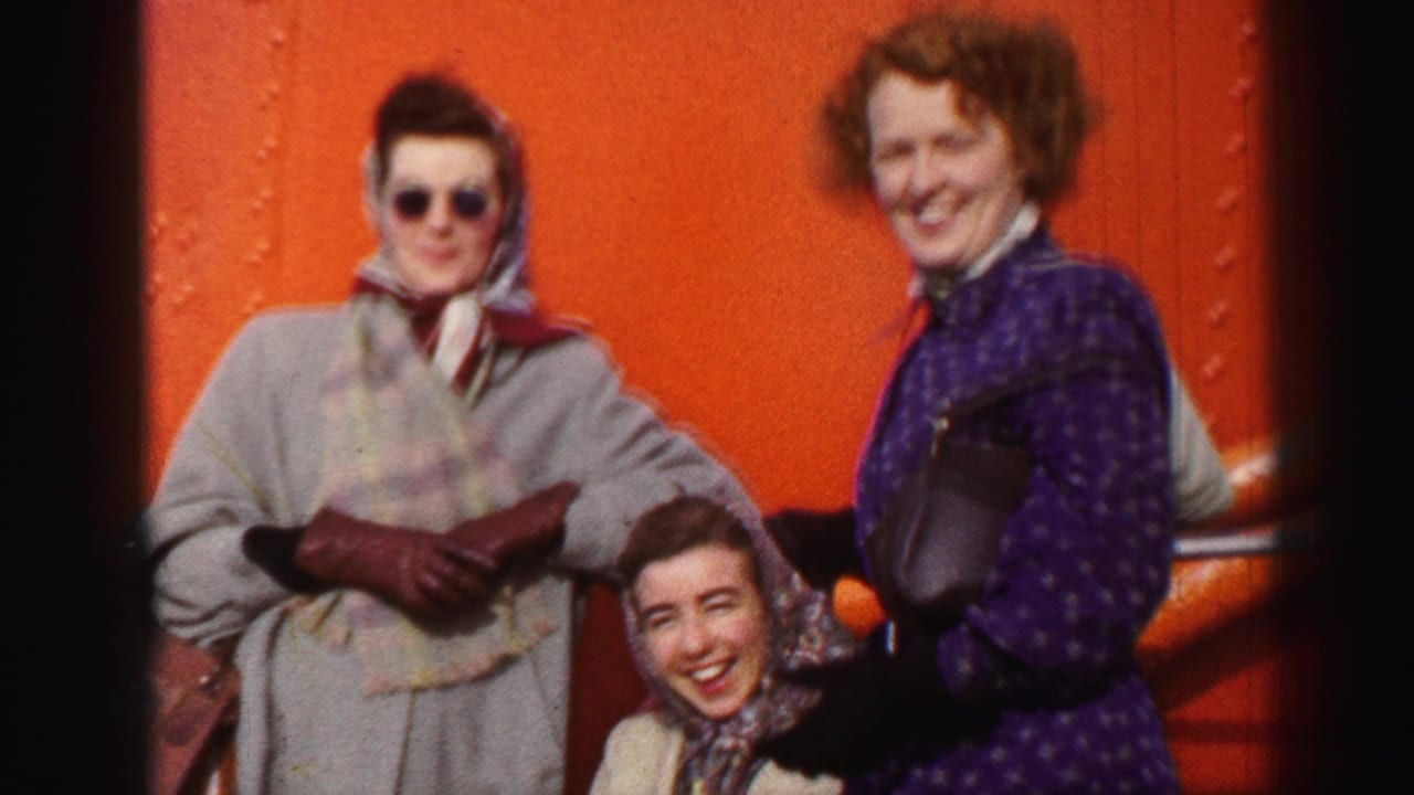 In supported 1952 United Kingdom home-movie footage, three women in mid-century coats, scarves, and gloves smile near an orange riveted ship-like wall.