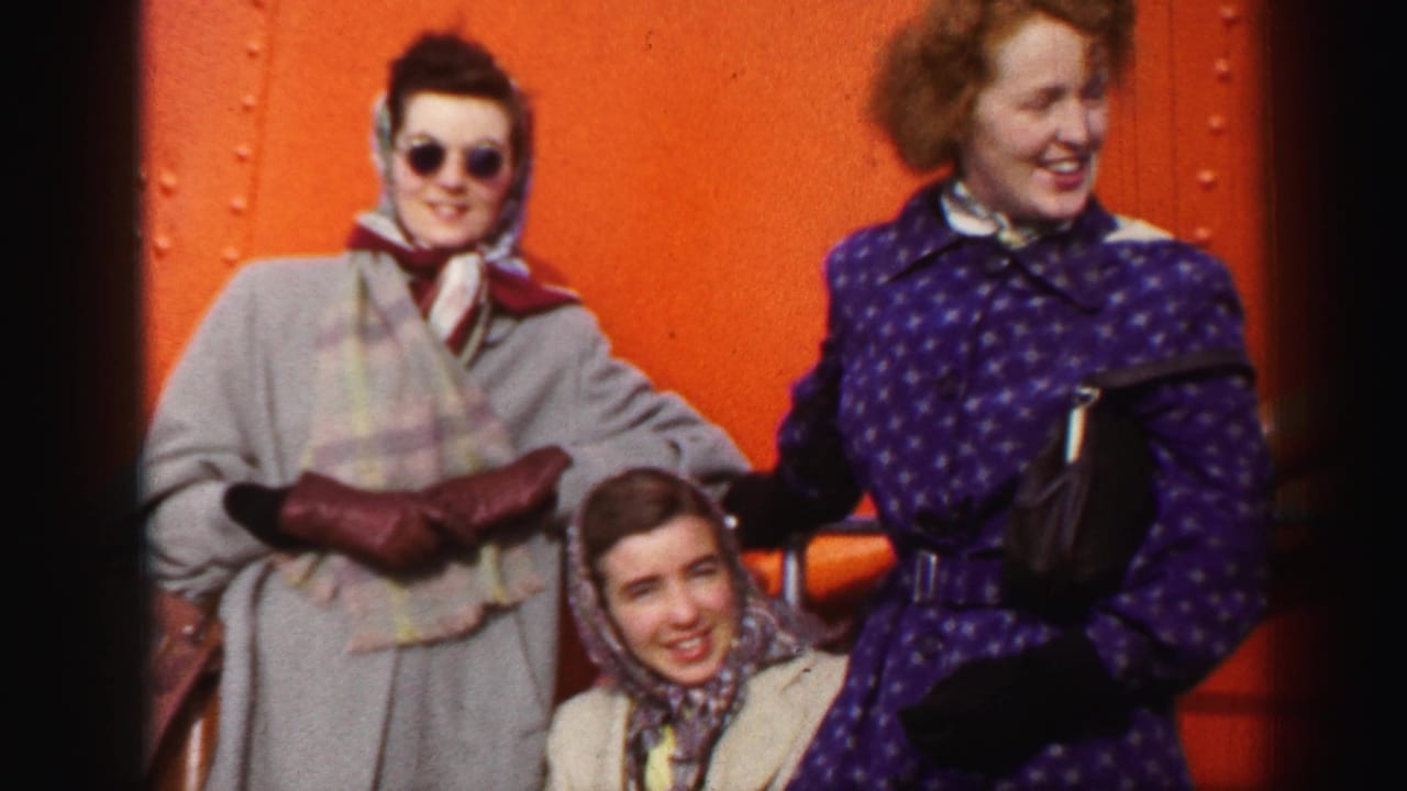 Three women in 1950s attire smile toward the camera beside a vivid orange riveted surface, likely on a ship in the United Kingdom footage.