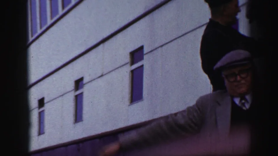 A brief handheld frame shows adults near a pale exterior wall with windows, suggesting movement around a building or access area after the posed shipboard shot.