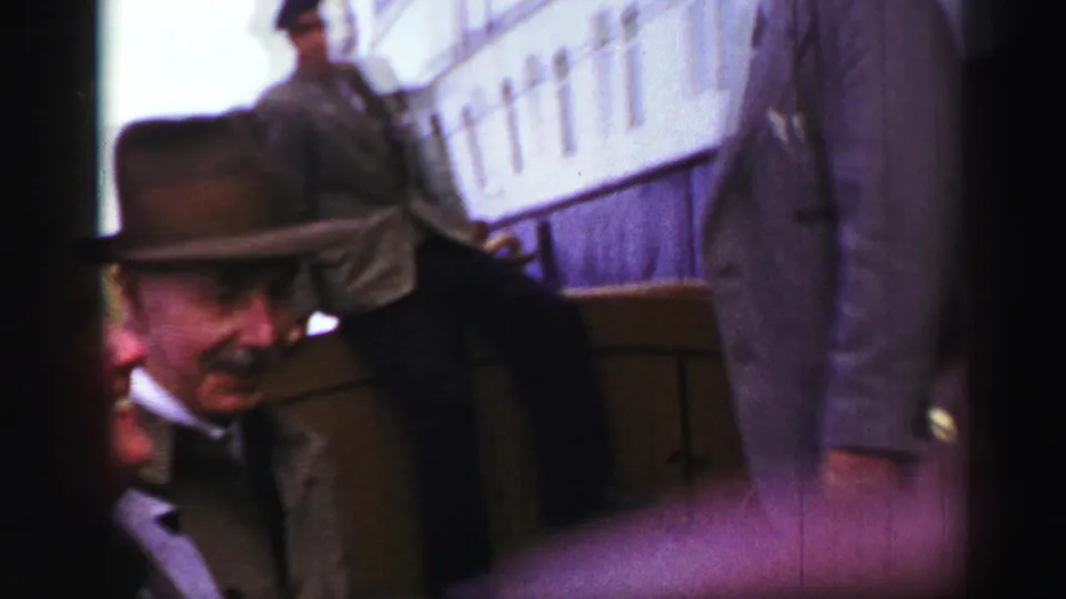 A blurred 8mm frame shows adult men near steps or railings, with a pale structure in the background and strong handheld motion.
