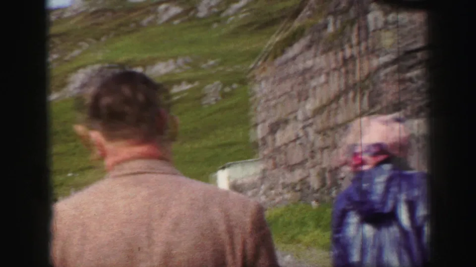 A man in a tan jacket walks through the ruin area while another visitor in a patterned coat stands near the stone wall.