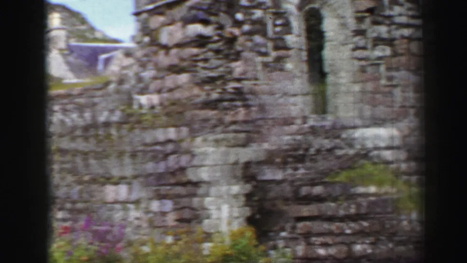Handheld 1952 United Kingdom frame of rough ruin walls, a narrow vertical opening, low masonry, and small plants in the foreground.