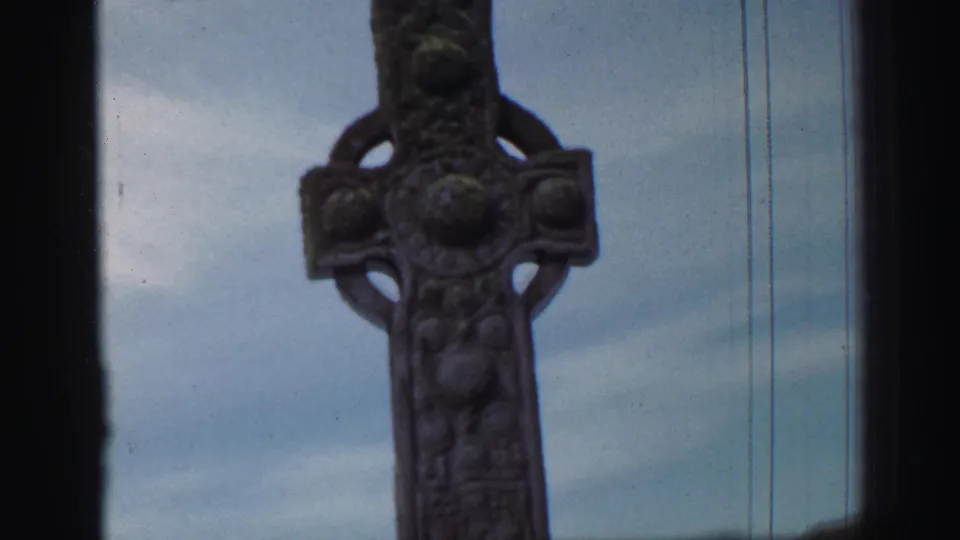 1952 United Kingdom frame showing a tall Celtic-style carved stone cross against cloudy sky with visible film scratches.