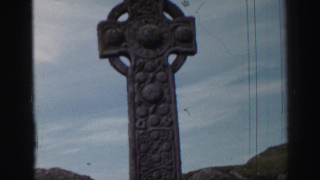 1952 United Kingdom 8mm frame showing a carved stone cross rising against cloudy sky with rocky ground behind it.