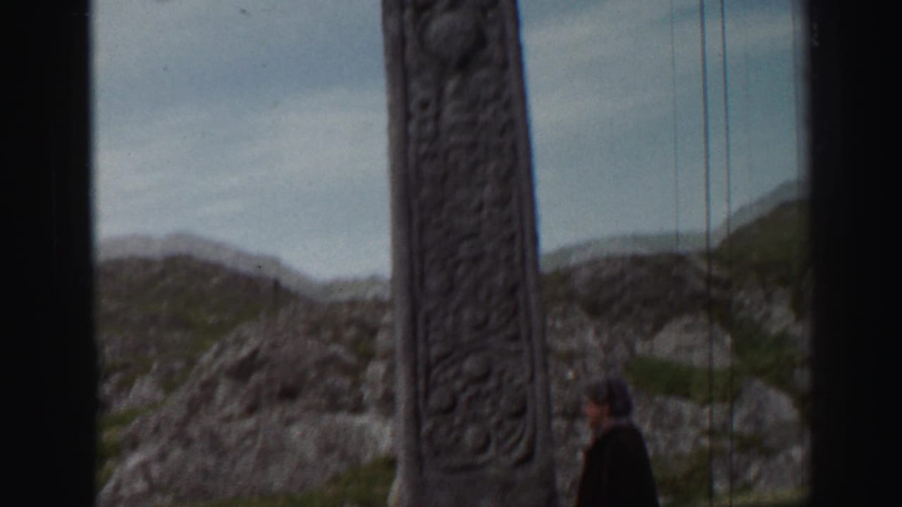Stone cross in foreground with a person walking near rocky terrain.