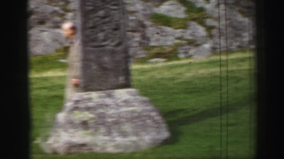 The camera frames the lower section and base of the carved stone cross set on grass below rocky terrain.