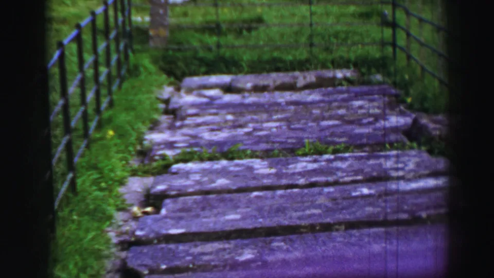 Close 1952 United Kingdom 8mm view of a weathered stone path, green grass, and black metal fencing near the church or abbey grounds.