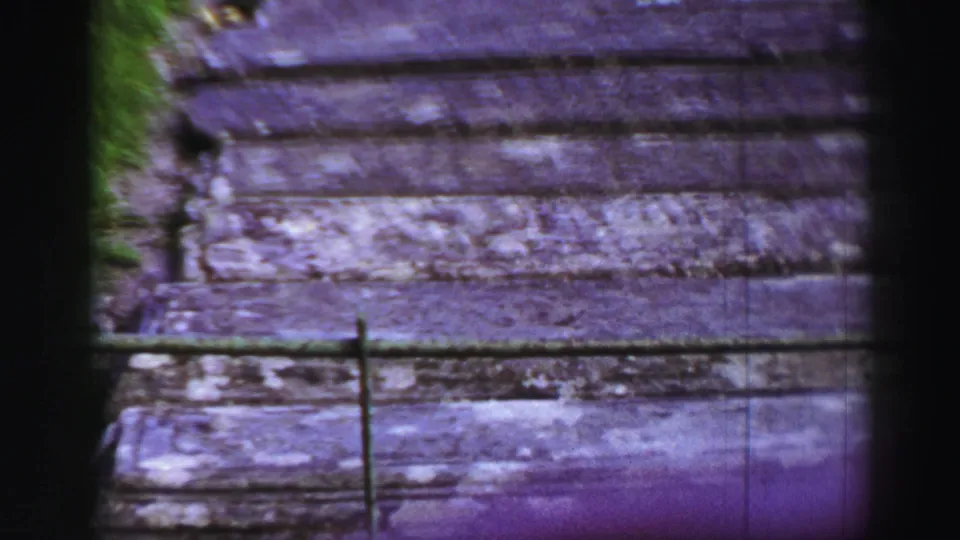 A handheld 8mm view looks along the weathered wooden path, with grass sides and railings cutting across the foreground.