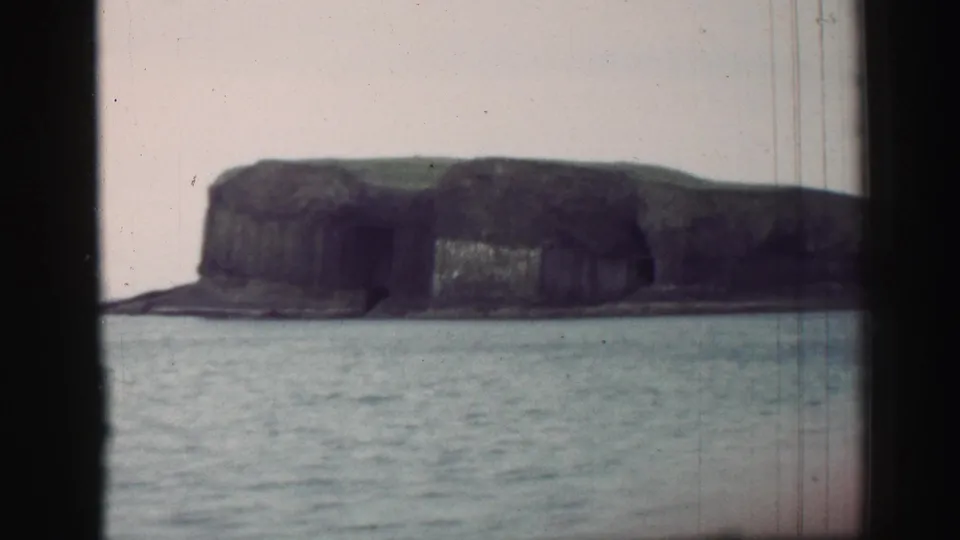 1952 United Kingdom 8mm view of a rugged cliffed island across water, seen through dark window or porthole framing.