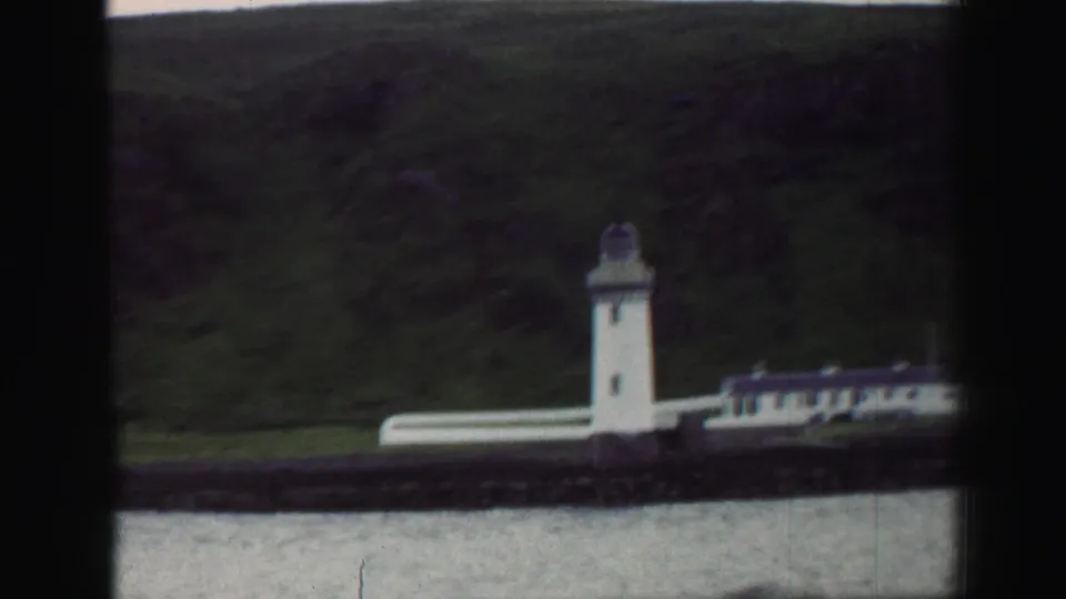 1952 United Kingdom 8mm frame with a white shoreline lighthouse, low buildings, water, and a dark hillside behind.