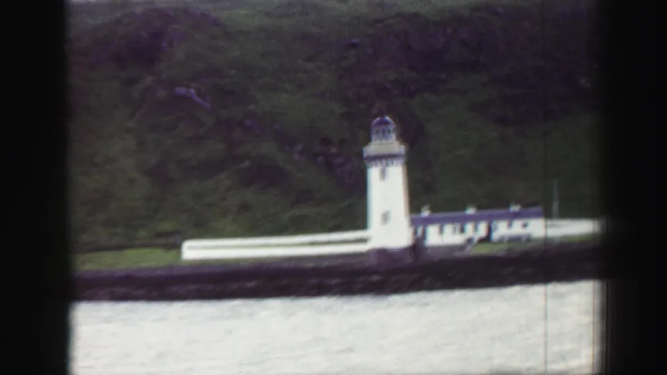 1952 United Kingdom coastal frame showing a white lighthouse complex beside water, backed by a grassy hillside.