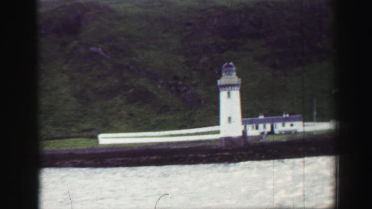 1952 United Kingdom 8mm frame showing a white lighthouse and adjacent buildings below a green hillside across the harbor.