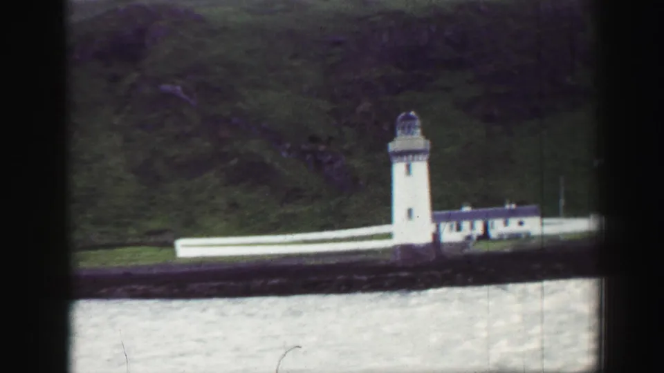 1952 United Kingdom 8mm frame showing a white lighthouse and adjacent buildings below a green hillside across the harbor.