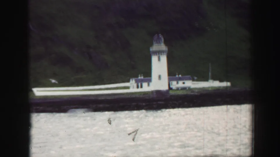 1952 United Kingdom harbor view with a white lighthouse, adjoining buildings, rocky seawall, and hillside behind.