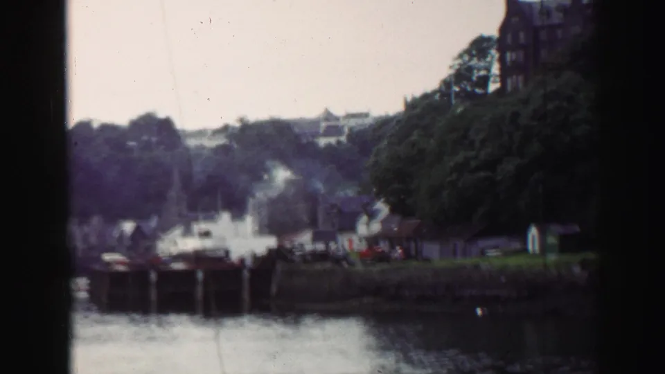 1952 United Kingdom 8mm frame showing a harbor village, docks, waterfront houses, trees, smoke, and hillside buildings.