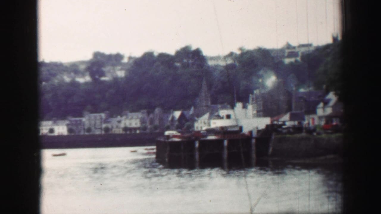 1952 United Kingdom harbour view with calm water, boats at a stone quay, distant buildings, and smoke rising near the waterfront.