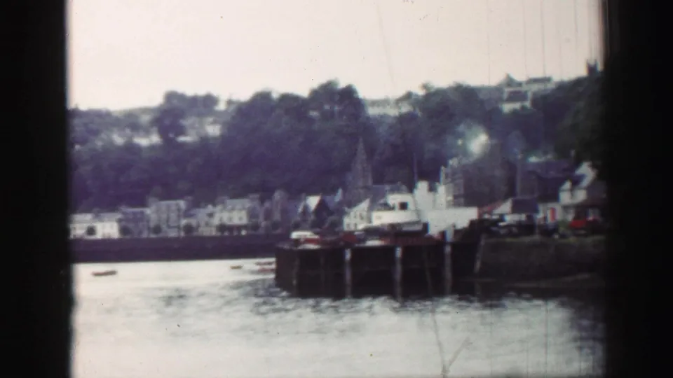 1952 United Kingdom harbour view with calm water, boats at a stone quay, distant buildings, and smoke rising near the waterfront.
