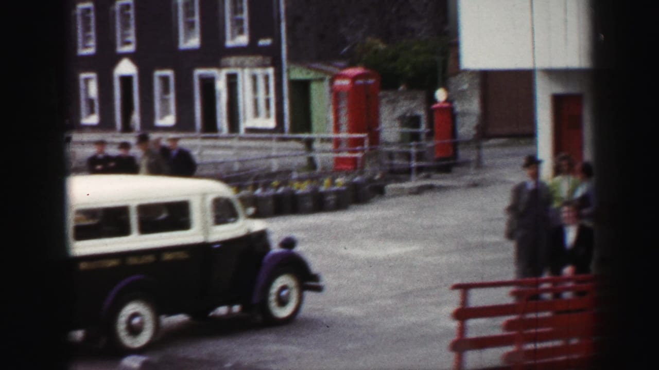 1952 United Kingdom street beside the harbour with a black-and-cream vehicle, red telephone boxes, waterfront buildings, and visible people.