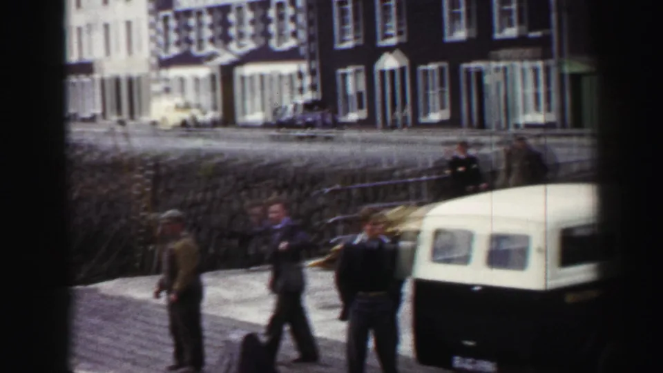 Men in period clothing stand and walk on a United Kingdom dockside in 1952, with a vehicle and waterfront buildings behind them.