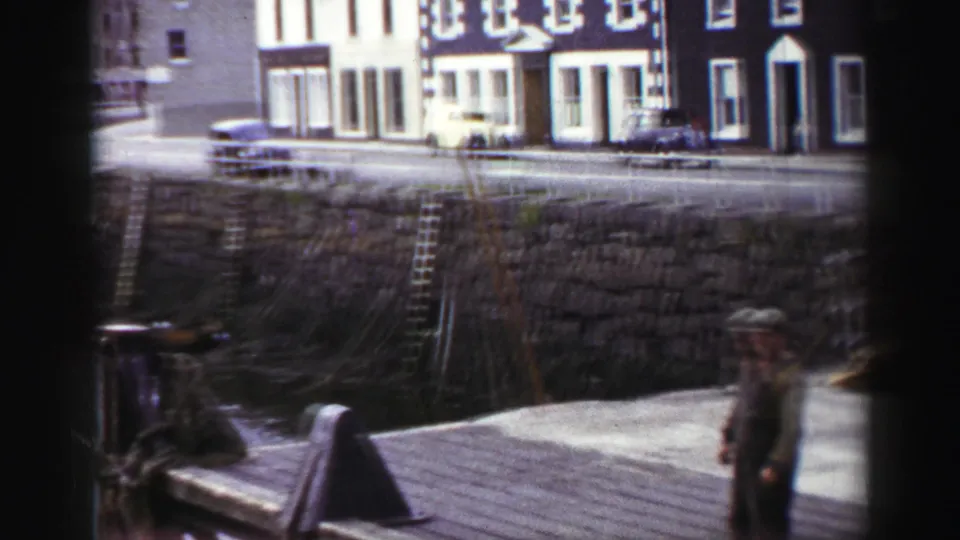 Stone quay wall and harbour street in the United Kingdom in 1952, with ladders, parked cars, waterfront buildings, and a man partly visible.