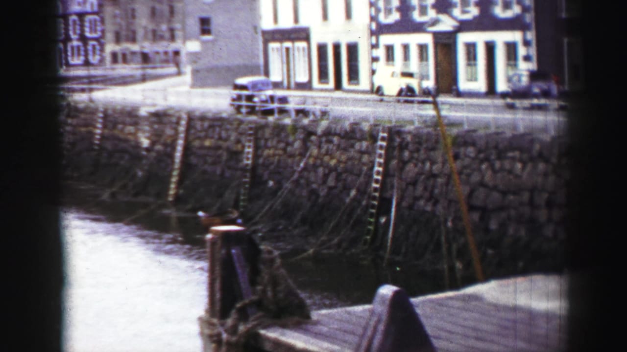 1950s United Kingdom home-movie view of a stone quay, wooden dock rails, parked car, and waterfront buildings, seen through soft 8mm framing.