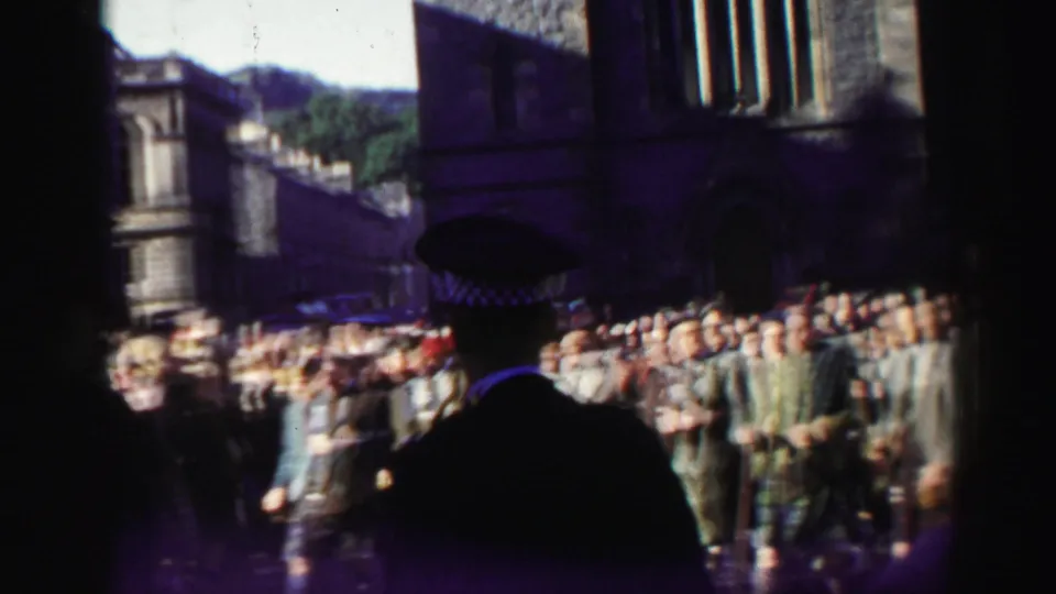 A uniformed police officer stands in silhouette before a crowd and kilted parade group, with dark stone buildings and hillside visible beyond.