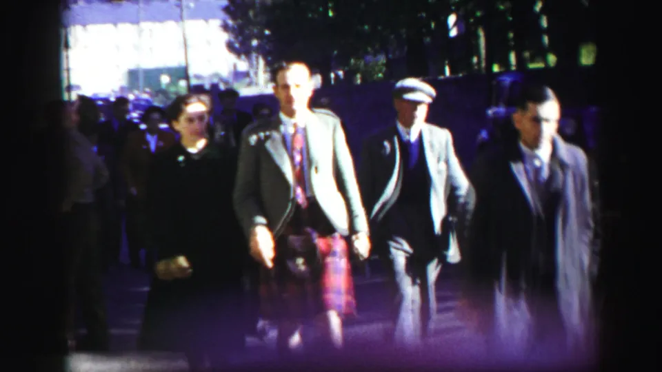 1952 United Kingdom home-movie frame of adults walking arm in arm on a city street, with men in kilts, tweed jackets, and women in period clothing.