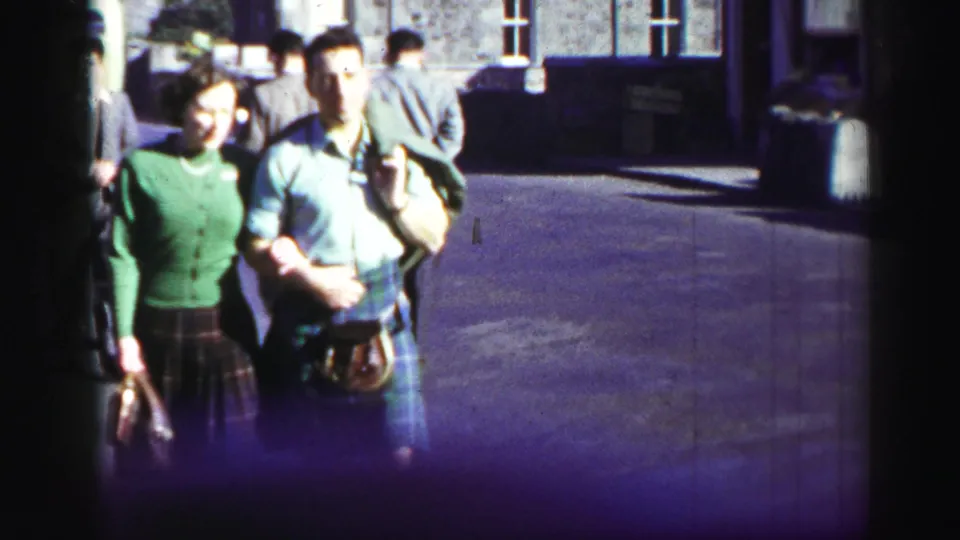 1952 United Kingdom home-movie frame of a woman in a green cardigan and plaid skirt walking arm in arm with a kilted man.