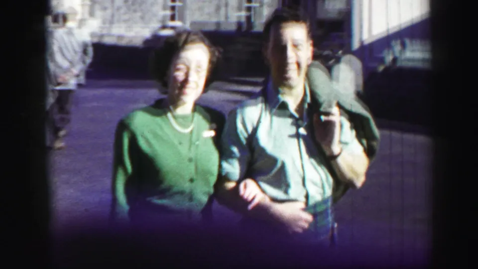 1952 United Kingdom frame of a smiling woman in a green cardigan beside a man in a kilt, with stone buildings and passersby behind them.