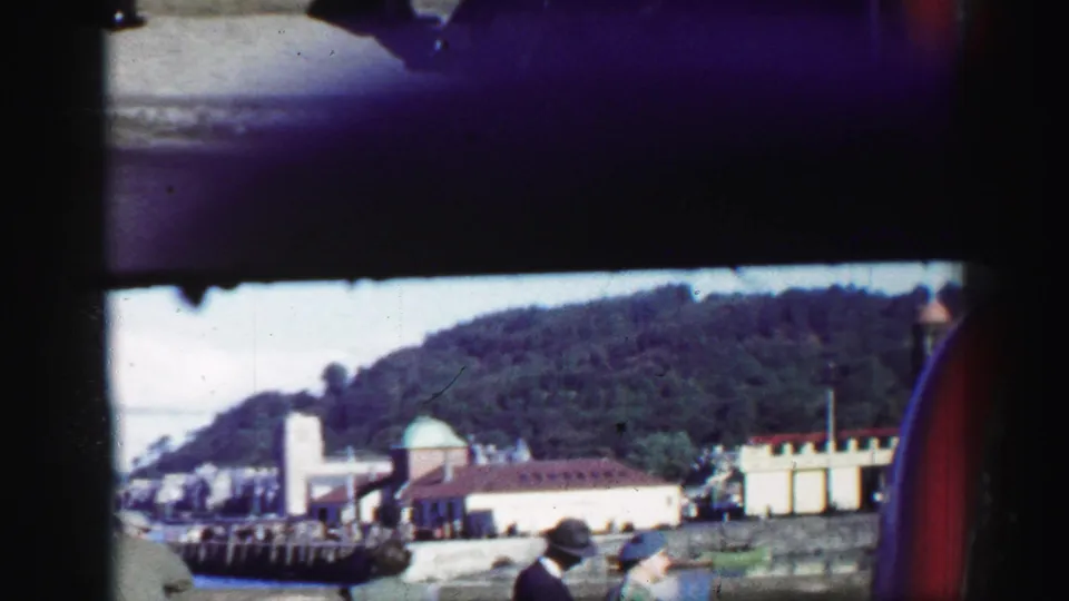 A harbor-side view shows coastal buildings, dock structures, water, and a wooded hillside in archival United Kingdom home-movie footage.