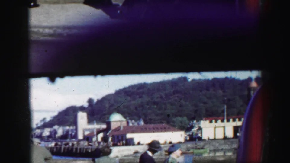 The coastal town view continues, showing harbor structures, pale waterfront buildings, wooded hillside, and a partial figure at the edge of frame.