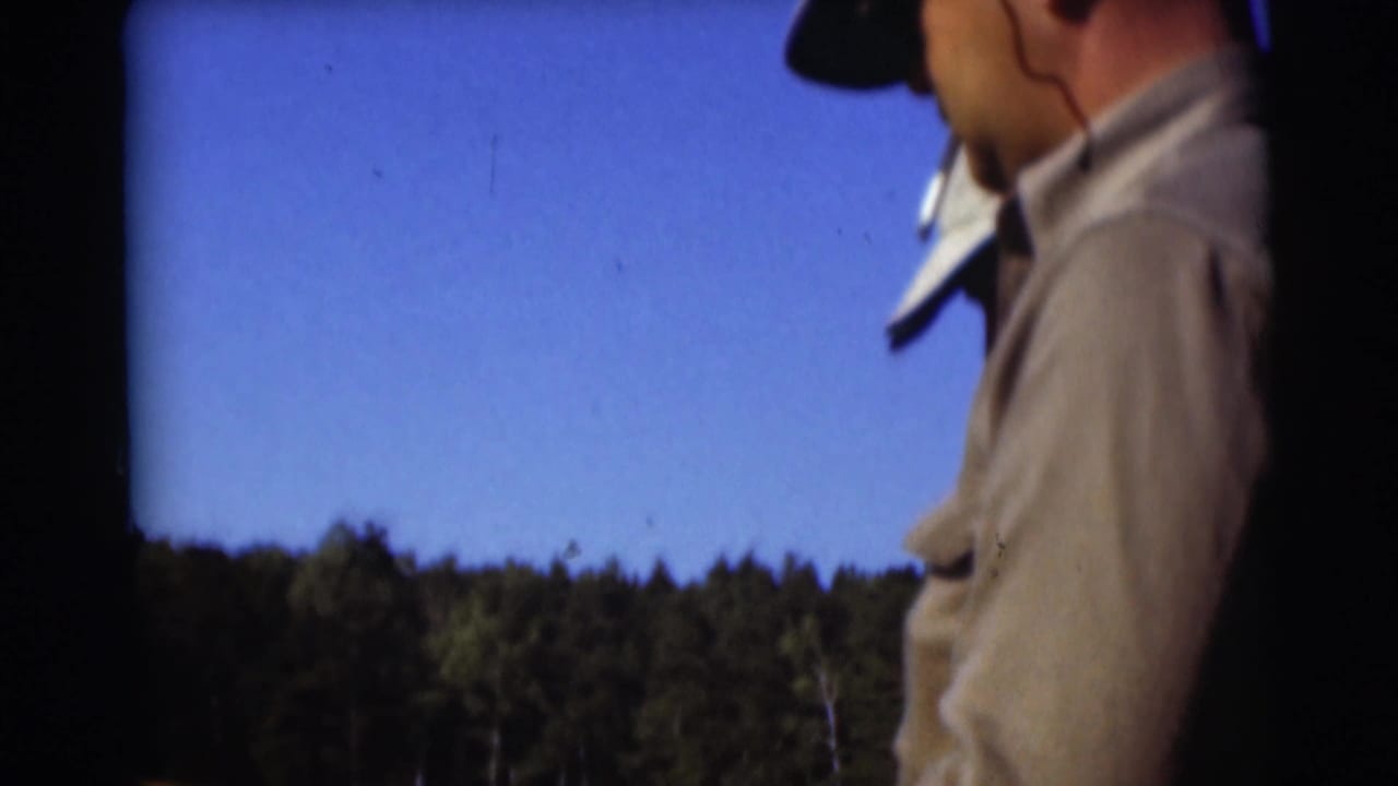A man in casual outdoor clothing stands near the lake, with a wooded shoreline and bright blue sky behind him.