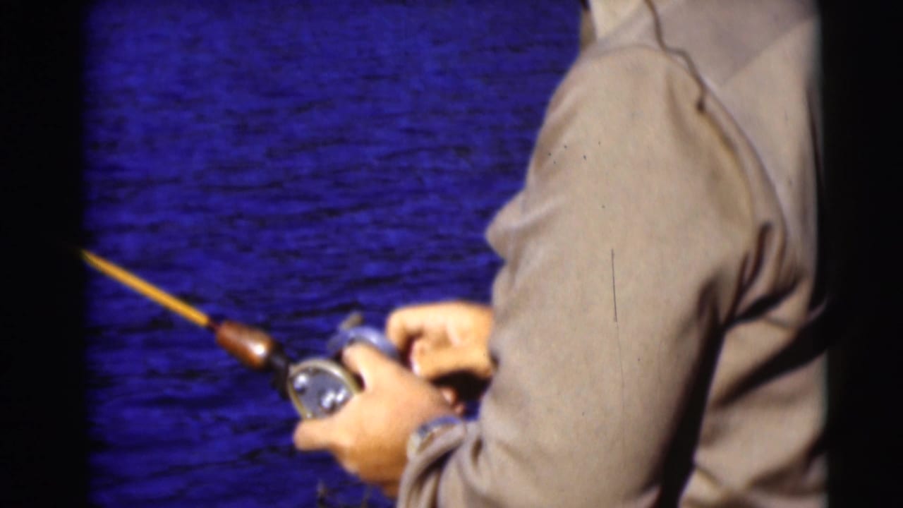 1953 Baraboo, Wisconsin home-movie frame of an adult angler reeling a fishing rod against a blue water backdrop.