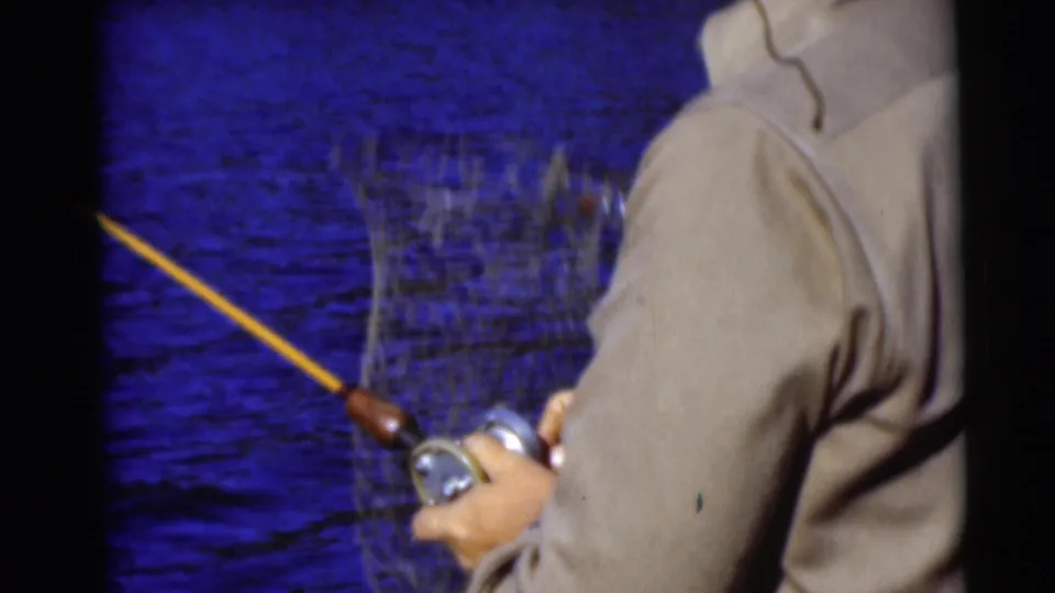 1953 Baraboo, Wisconsin frame with an adult angler, rod, reel, and landing net visible during a fishing moment on the water.