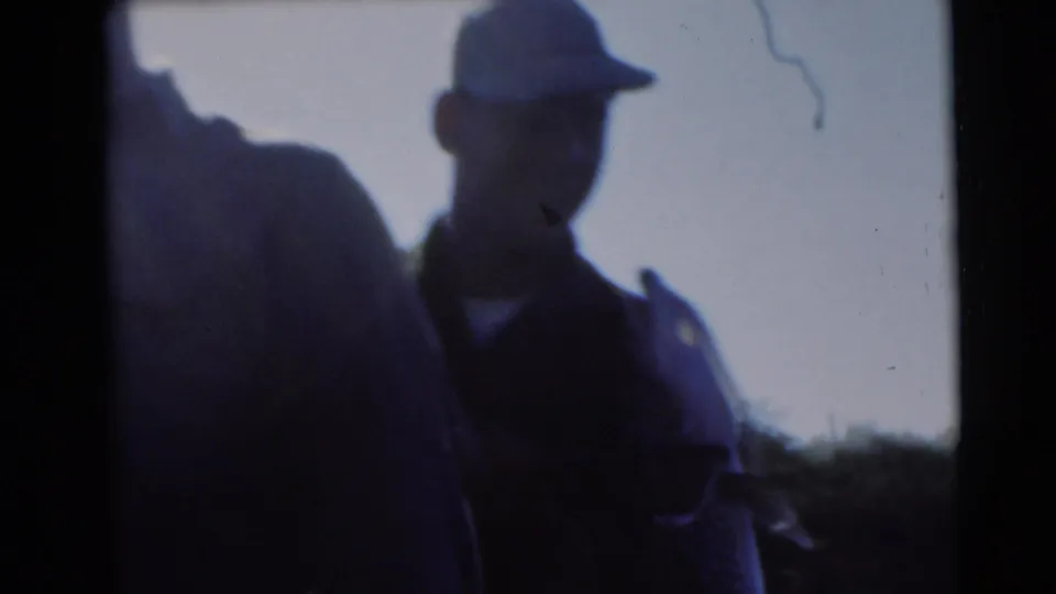 A capped man stands in silhouette beside the upright northern pike, with lake edge and trees faintly visible in the 8mm frame.