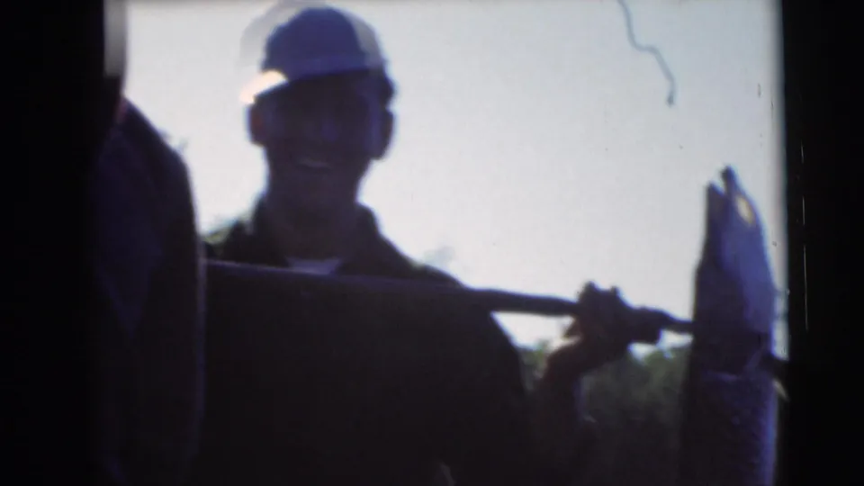 Man in a cap smiles while holding a pole with a large fish in a 1953 Baraboo, Wisconsin home movie.