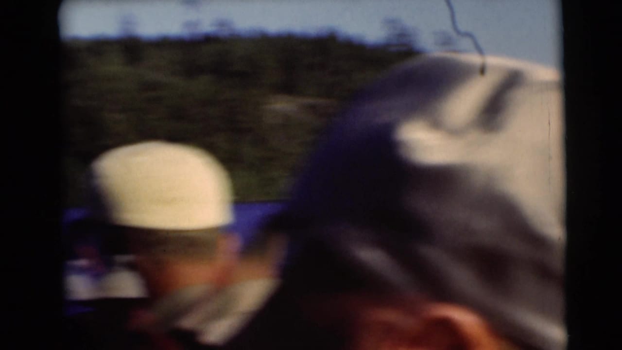 Soft 1953 home-movie frame from Baraboo showing two capped men on a boat, with lake water and a tree-lined shore in the background.