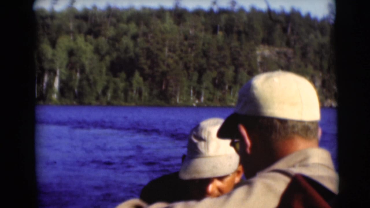 Two men in caps sit near the camera on a boat with blue water and forest behind them.