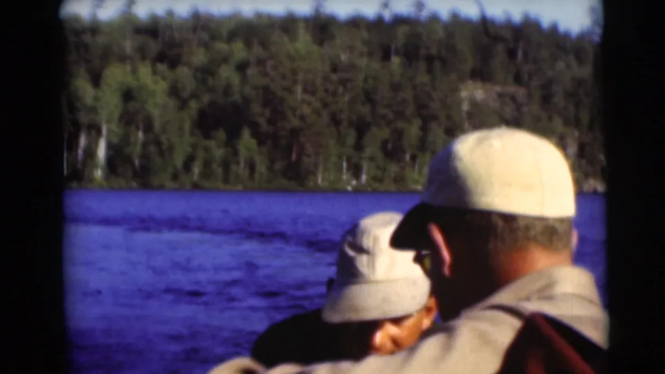 Two men in beige caps appear aboard a boat on a Baraboo lake in 1953, with blue water and dense trees across the shoreline.