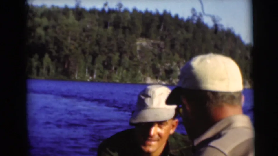 A 1953 Baraboo lake frame shows two men in light caps on a boat, one facing the camera, against blue water and a wooded shoreline.