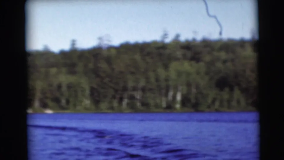 The camera looks across a Baraboo lake toward a dense green shoreline, showing water, trees, sky, and soft 8mm film texture.