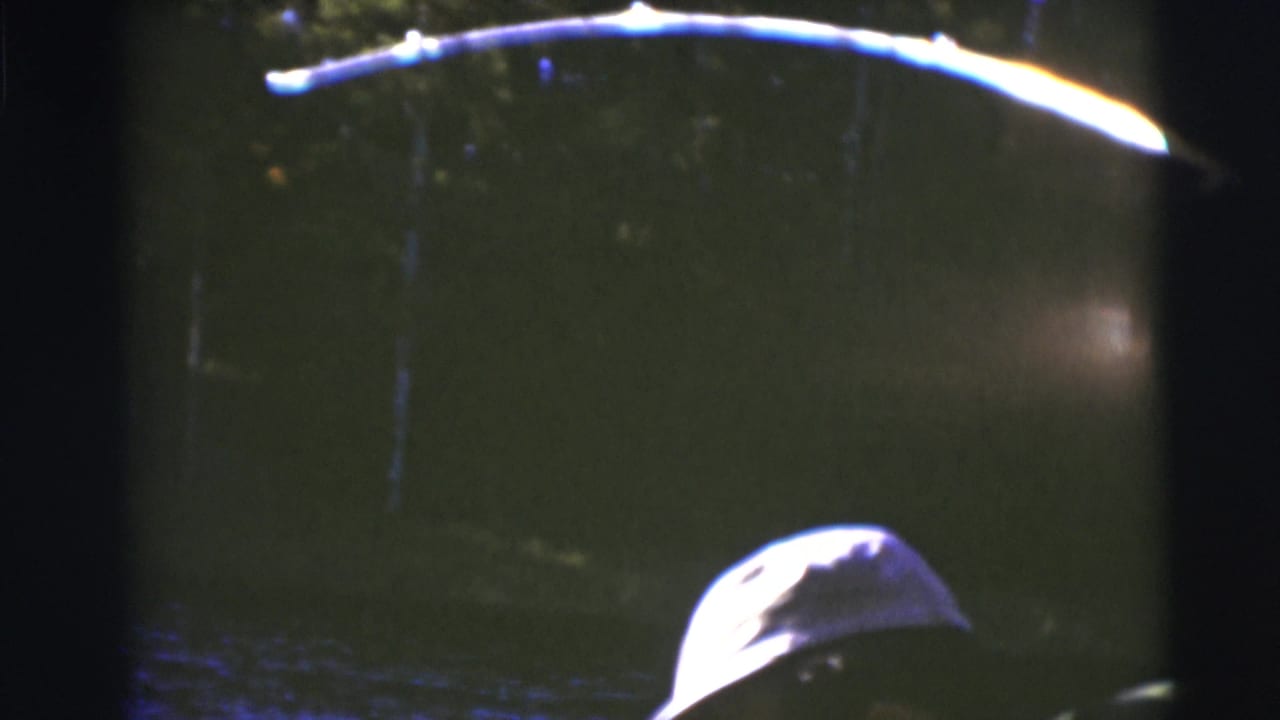 1953 Baraboo home-movie frame showing dark lake water, a bright curved reflection or pole shape, and part of a capped head at the edge.