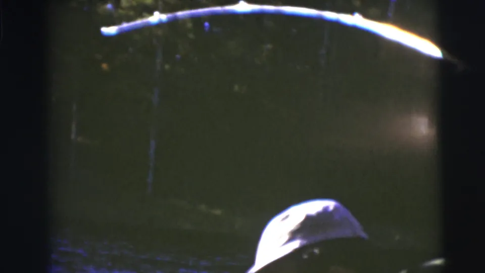 1953 Baraboo home-movie frame showing dark lake water, a bright curved reflection or pole shape, and part of a capped head at the edge.