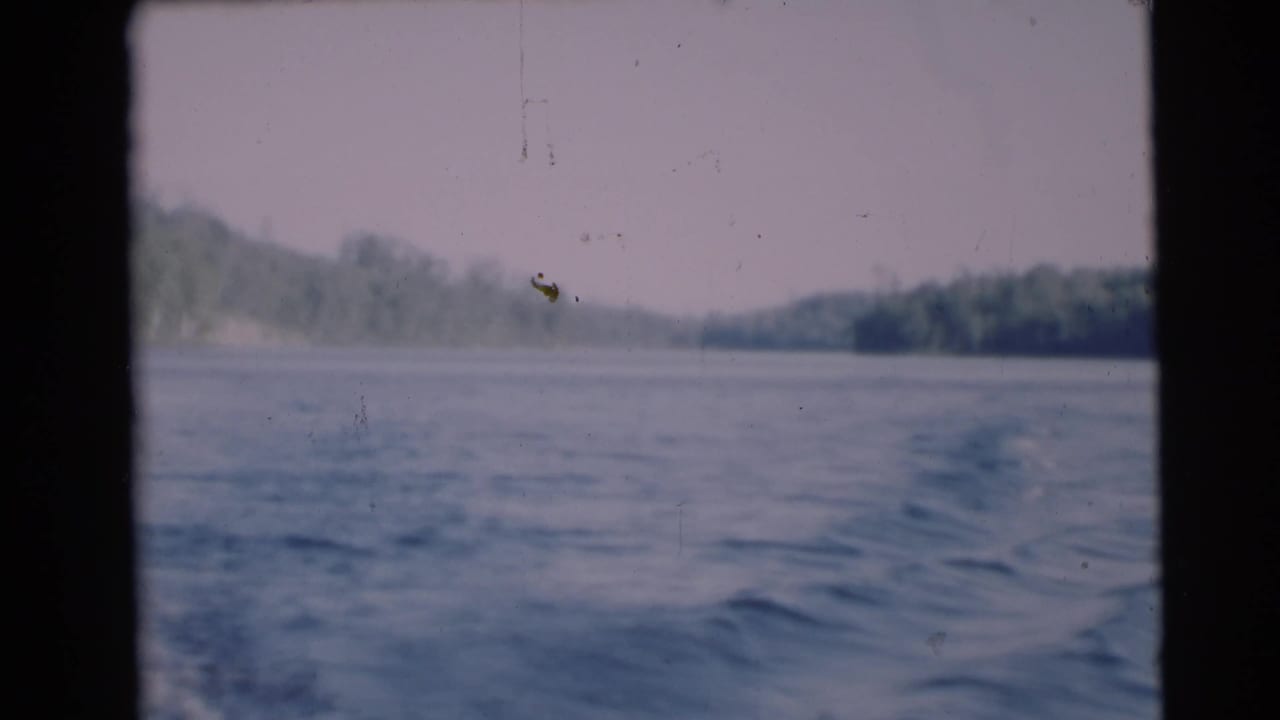 1953 8mm view from a moving boat near Baraboo, Wisconsin, showing rippling water, pale sky, and wooded shoreline.